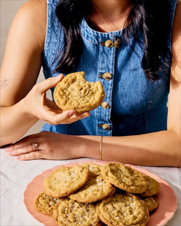 A baker holding a brown butter cookie above a plate of cookies.