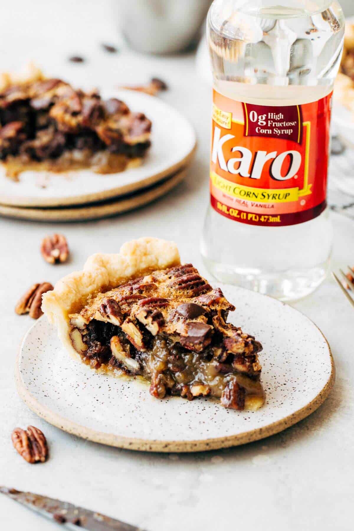 A slice of chocolate pecan pie on a white plate with a bottle of corn syrup in the background.