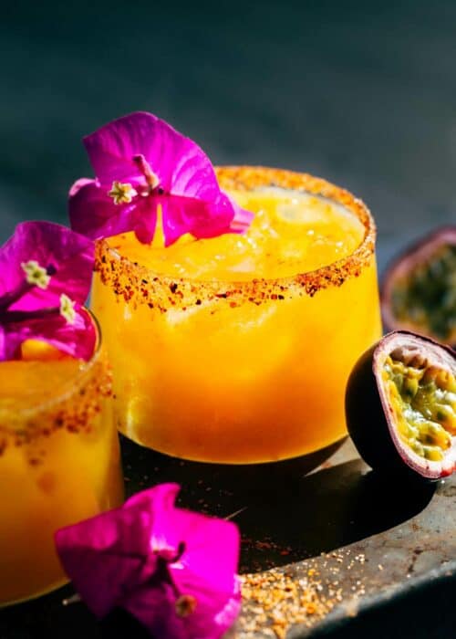Two spicy passion fruit margaritas surrounded by decorative flowers and half of a passion fruit
