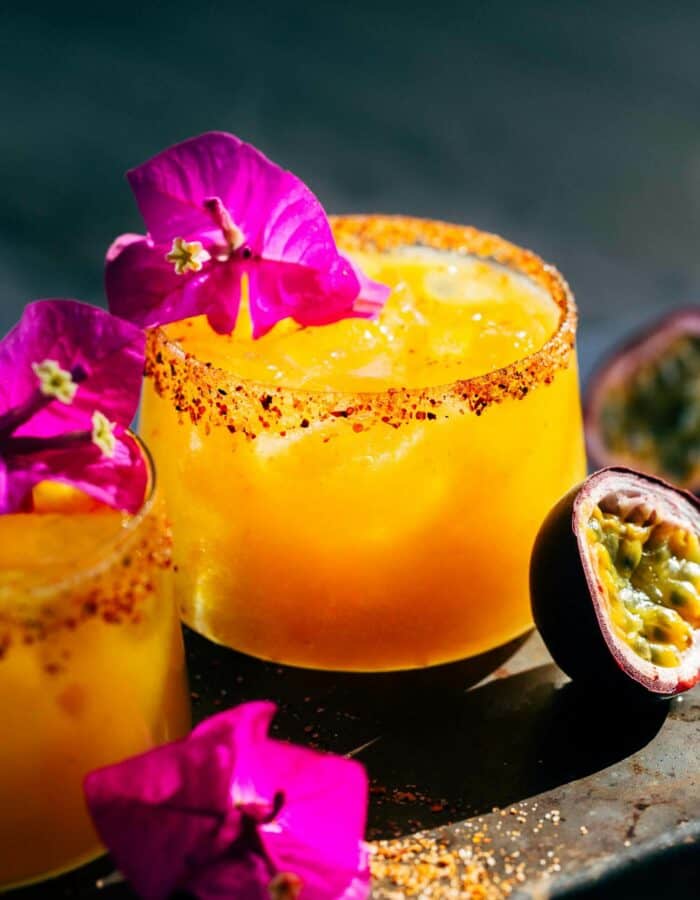 Two spicy passion fruit margaritas surrounded by decorative flowers and half of a passion fruit